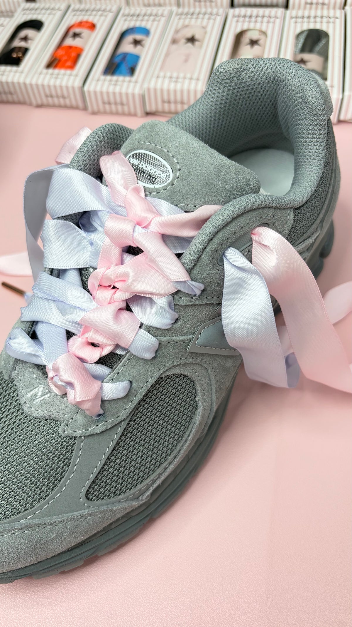 A gray mesh sneaker with decorative pastel pink and light blue ribbon laces, photographed on a pink surface with small product boxes blurred in the background.